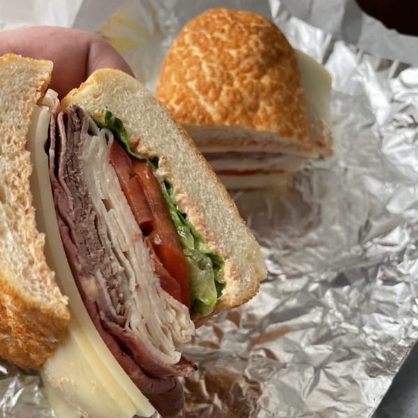A hand holds a deli sandwich with roast beef, turkey, cheese, lettuce, and tomato, with another sandwich in foil behind it.