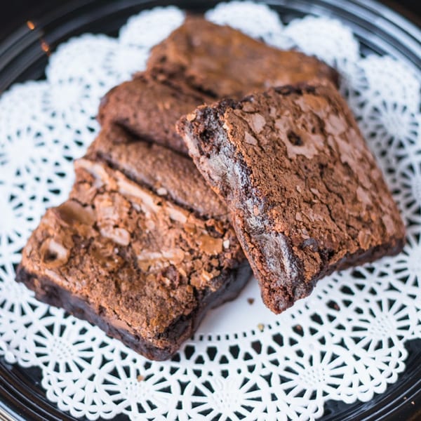 Several rich, fudgy brownies with crinkled tops on a white doily on a black plate.