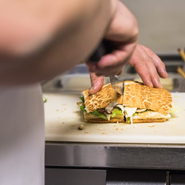 Chef's hands slicing a toasted deli sandwich with lettuce and cheese on a cutting board.