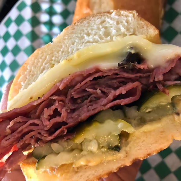 Close-up of a deli sandwich with pastrami, melted cheese, and pickles on a bun.